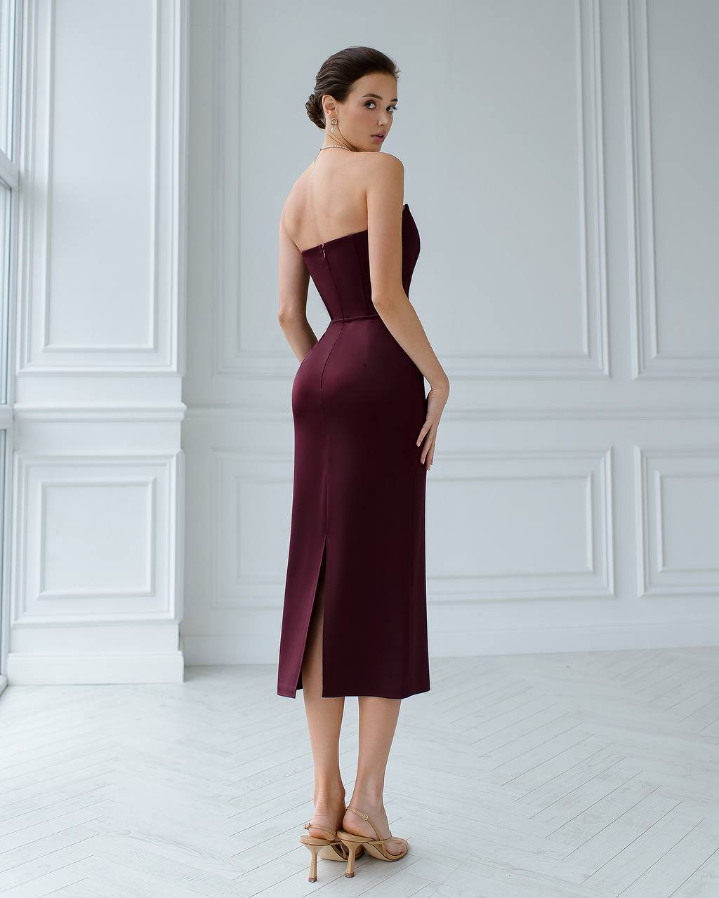 Woman wearing a burgundy strapless dress with a slit in a white room.