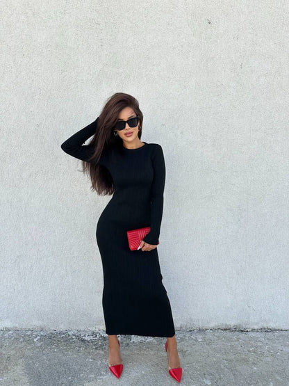 Woman in a black dress with red shoes and clutch against a white wall