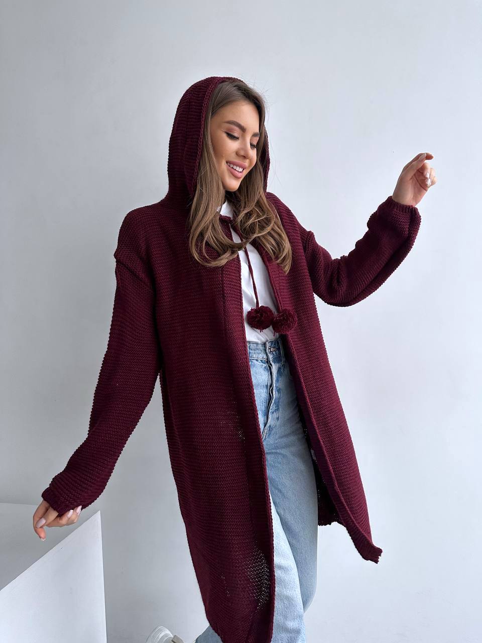 Woman wearing a burgundy hooded coat with pom-pom details on a plain background