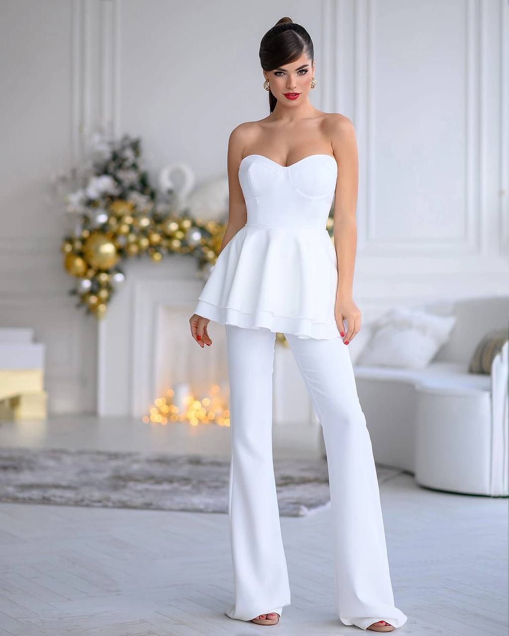 Woman wearing a white strapless top and flared pants in a decorated room.