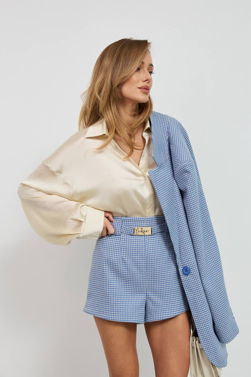 Woman wearing a beige blouse, blue checkered shorts, and a blue blazer against a white background