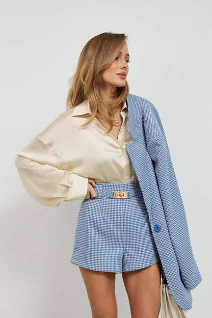 Woman wearing a beige blouse, blue checkered shorts, and a blue blazer against a white background