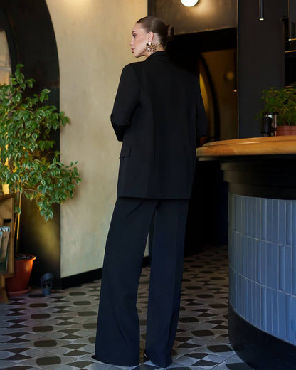 Person wearing a black suit standing in a room with plants and a counter.
