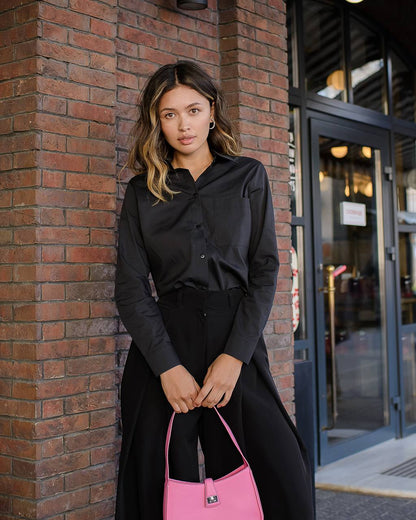Woman in black outfit holding a pink handbag against a brick wall.