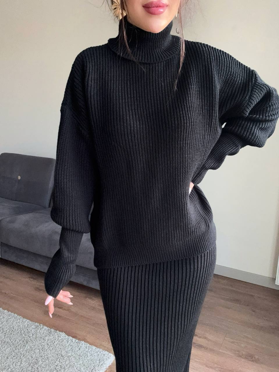 Person wearing a black ribbed sweater and skirt set in a room with a gray couch.