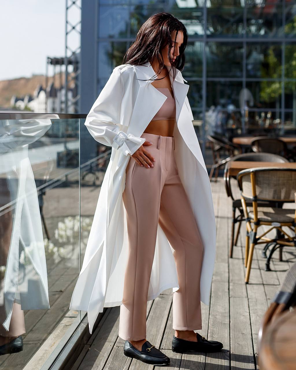 Woman in a white trench coat and pink outfit standing on a wooden deck with a cityscape background.