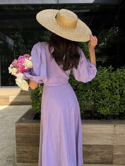 Woman in a lavender dress and straw hat holding flowers, standing outdoors.