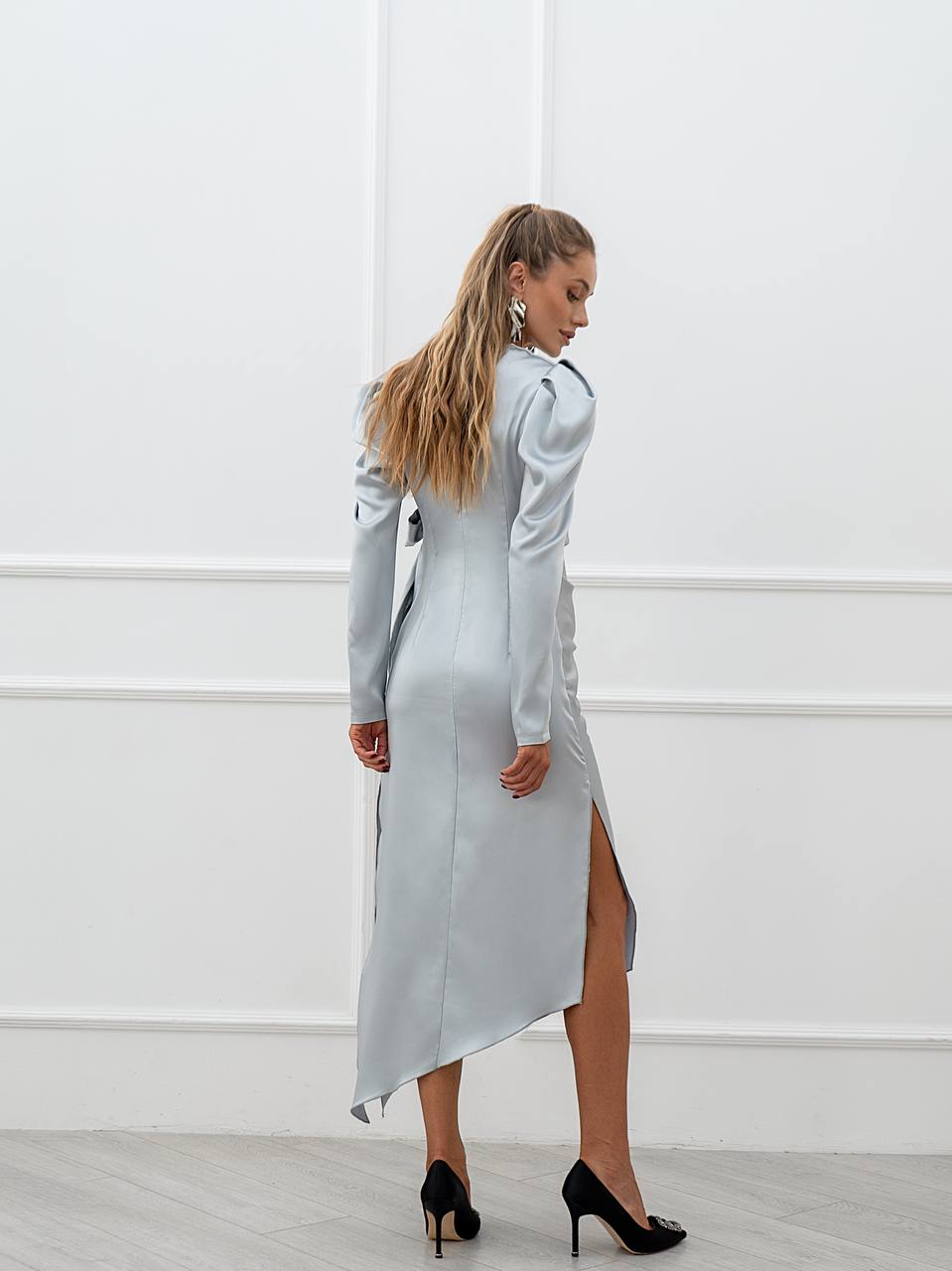 Woman wearing a light gray dress with a high slit, standing against a white wall.