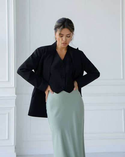 Woman wearing a black blazer and light green skirt against a white paneled wall.