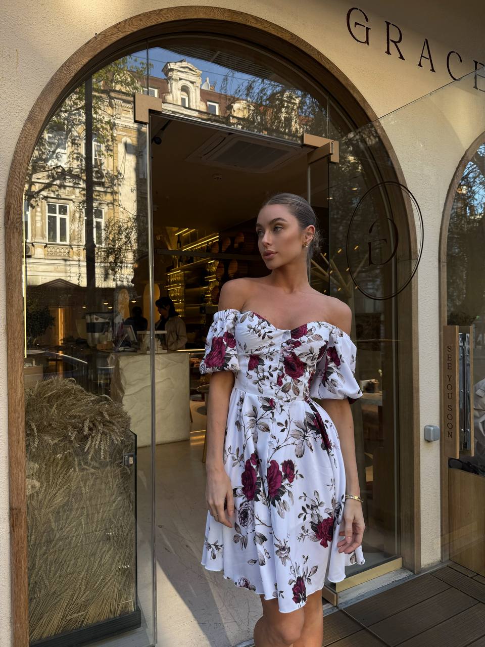 Woman in a floral off-shoulder dress standing in front of a store named 'Grace'.