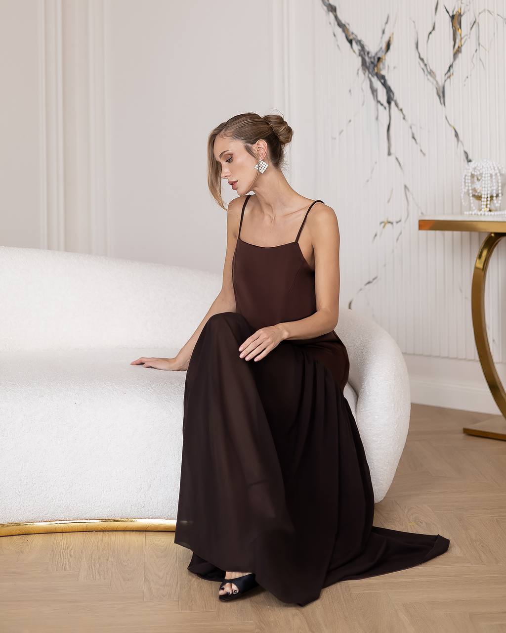 Woman in a long brown dress sitting on a white chair in a stylish room.