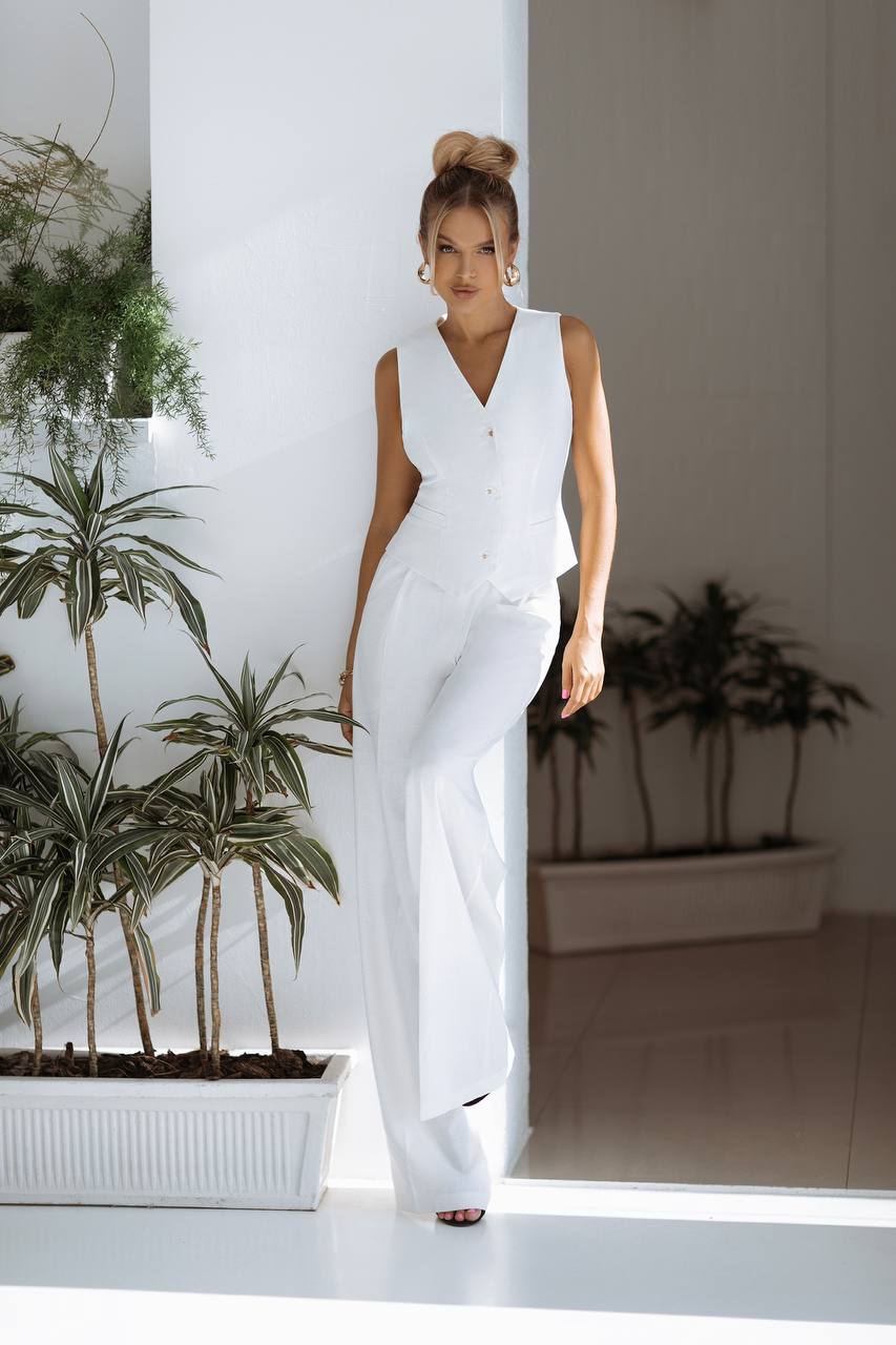 Woman in a white sleeveless top and pants standing in a modern indoor setting with plants.