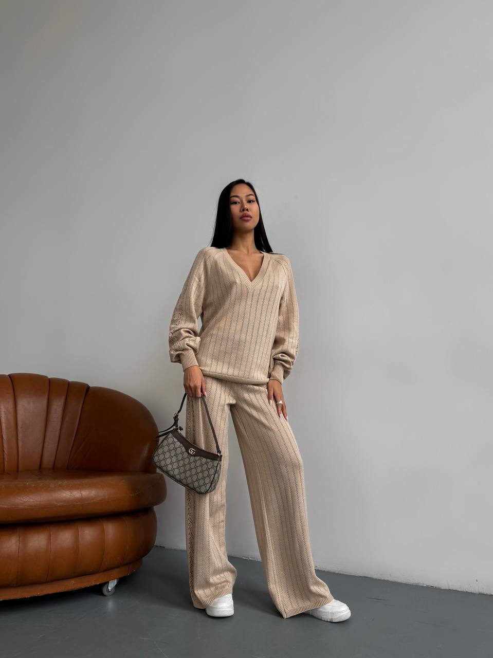 Woman in a beige outfit standing next to a brown leather chair against a gray wall.