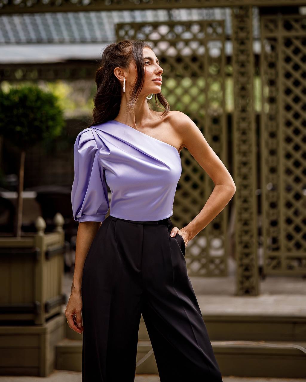 Woman wearing a one-shoulder lavender top and black pants outdoors.