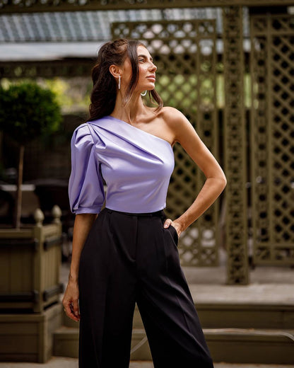 Woman wearing a one-shoulder lavender top and black pants outdoors.