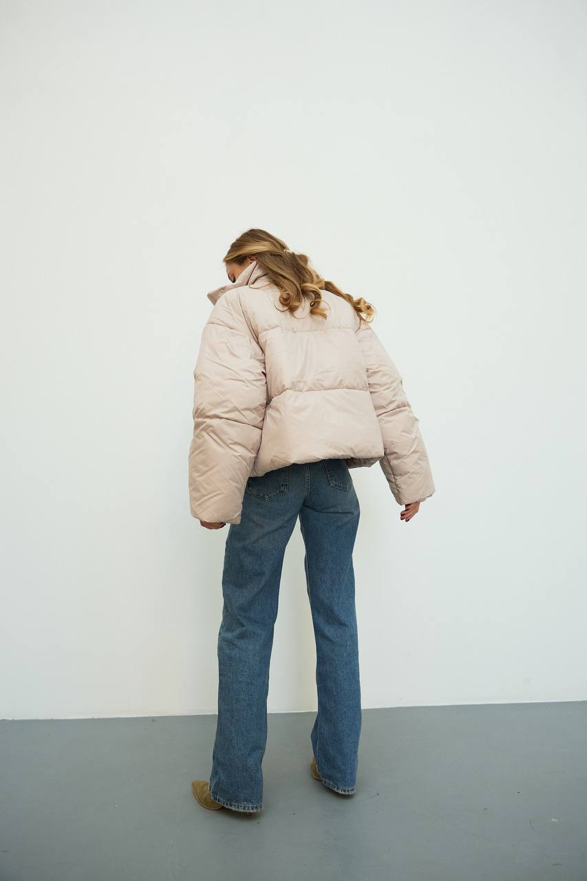 Person wearing a beige puffer jacket and blue jeans standing against a white wall.