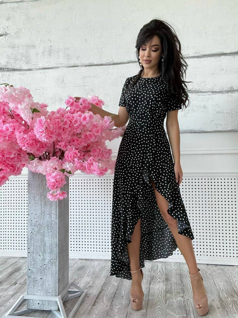 Woman in a black polka dot dress standing next to pink flowers indoors.
