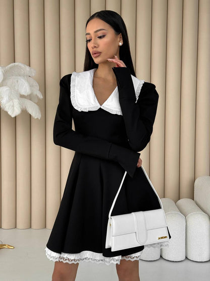 Woman wearing a black dress with white accents in a room with beige curtains and a white chair.