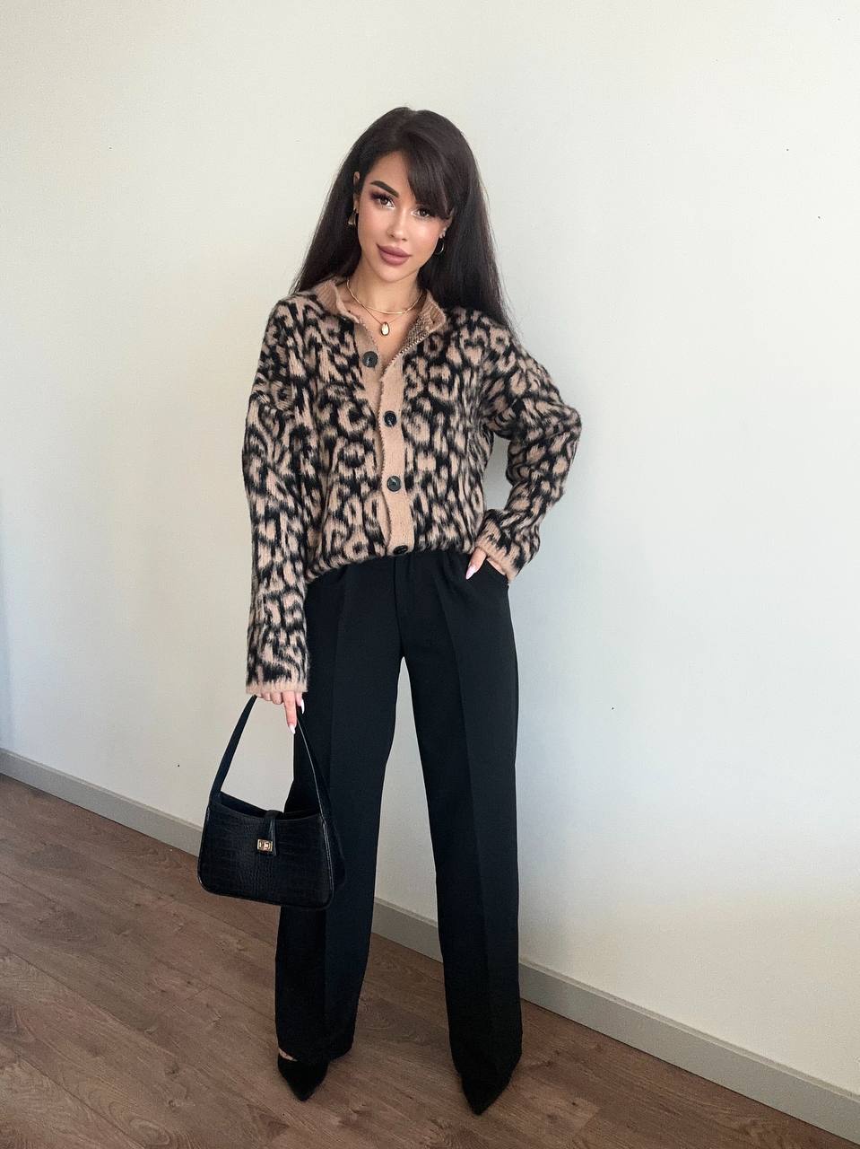 Woman wearing a leopard print jacket and black pants, standing against a white wall.
