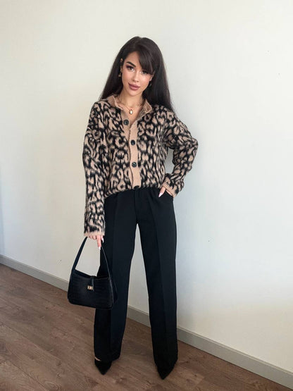 Woman wearing a leopard print jacket and black pants, standing against a white wall.