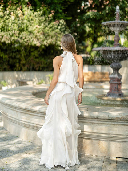 A woman wearing a flowing white halter-neck maxi dress with layered ruffles, photographed from behind in an outdoor garden setting with a stone fountain and greenery in the background.