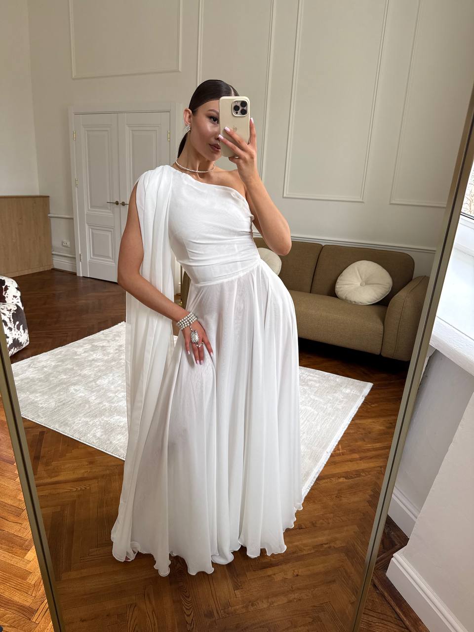 Woman in a white dress taking a mirror selfie in a room with wooden floor and beige sofa.