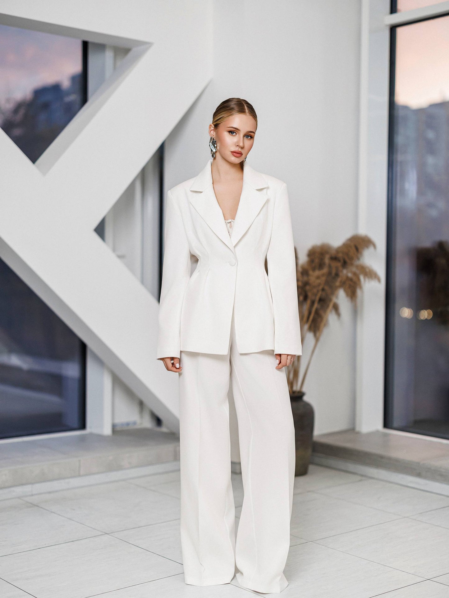 Woman in a white suit standing in a modern indoor setting with large windows.