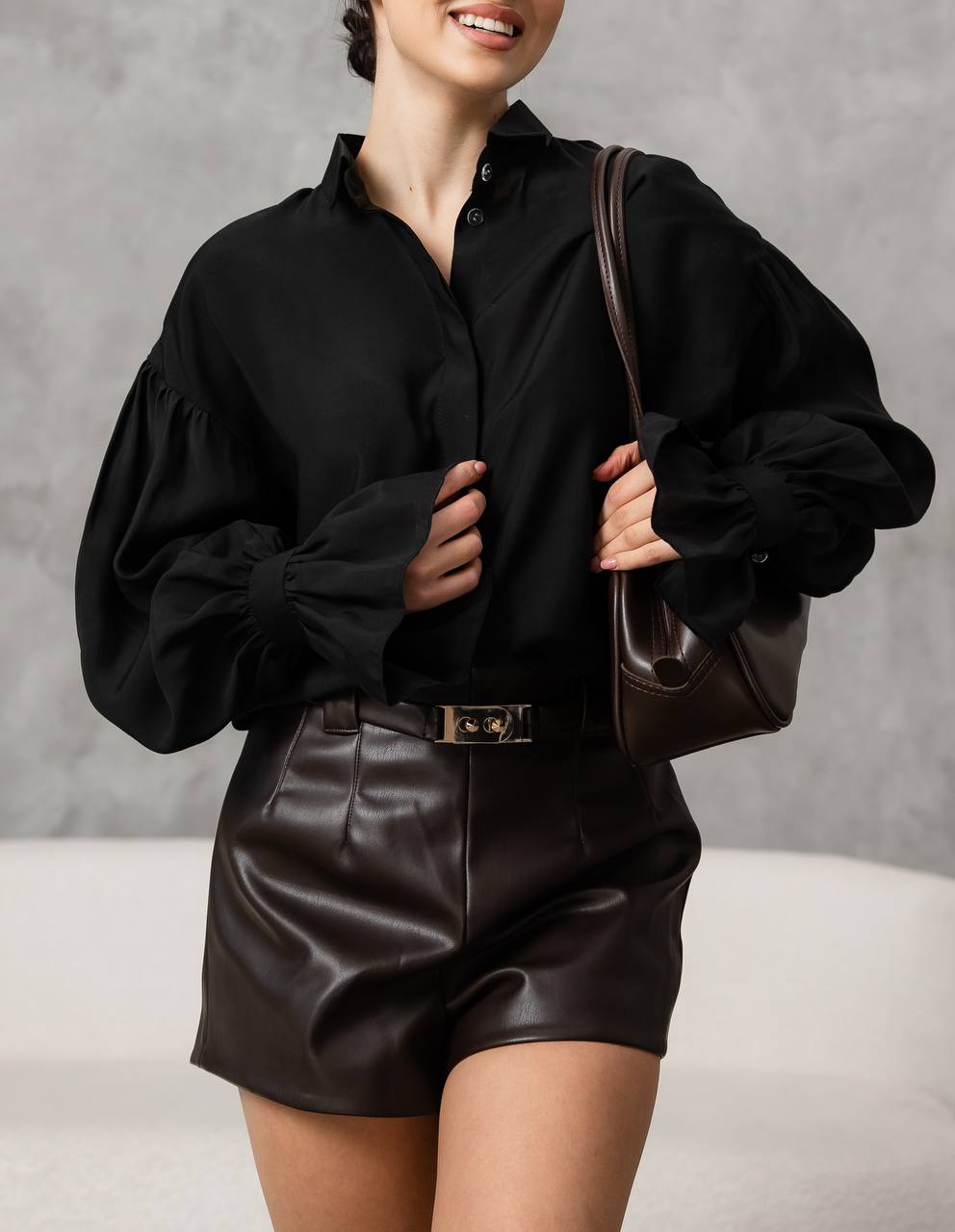 Person wearing a black shirt and leather shorts holding a brown bag against a gray background