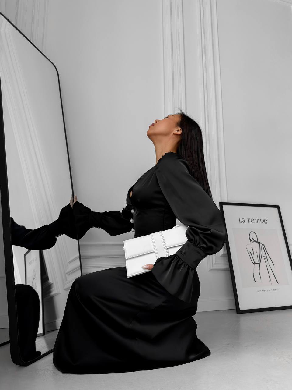 Woman in a black dress holding a white clutch in front of a mirror.
