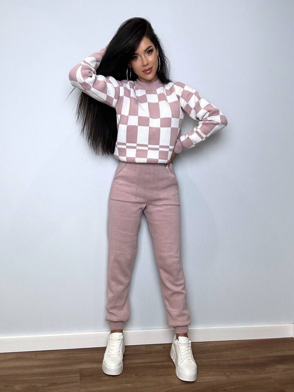 Woman wearing a checkered pink and white sweater and matching pants against a plain wall.