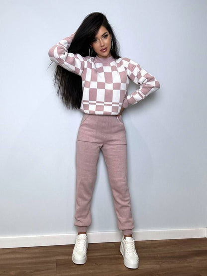 Woman wearing a checkered pink and white sweater and matching pants against a plain wall.