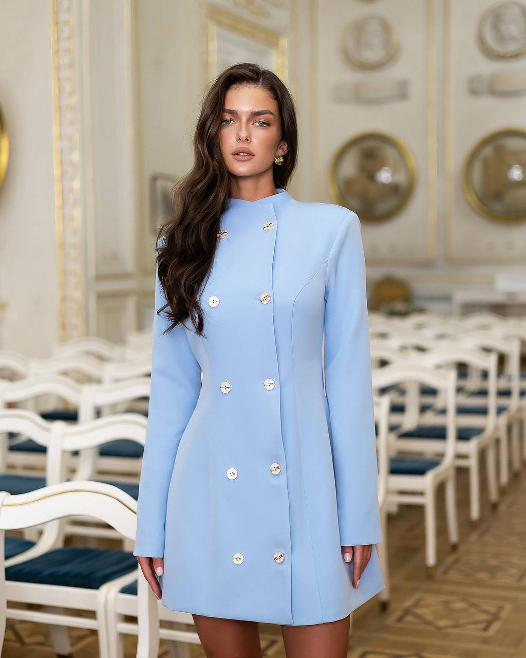 Woman wearing a light blue coat with gold buttons in an elegant interior setting.