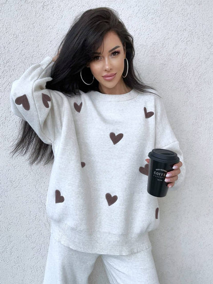 Woman wearing a white sweater with brown heart patterns, holding a black coffee cup against a plain background.