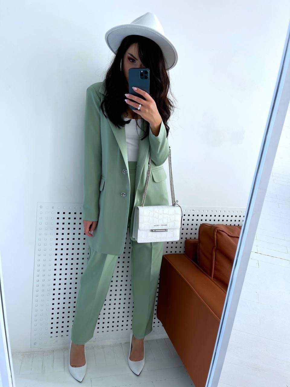 Person wearing a green suit and white hat, holding a phone, with a white wall and brown couch in the background.