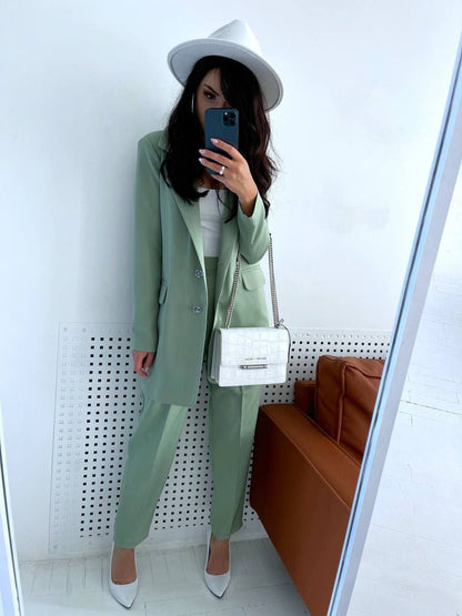 Person wearing a green suit and white hat, holding a phone, with a white wall and brown couch in the background.