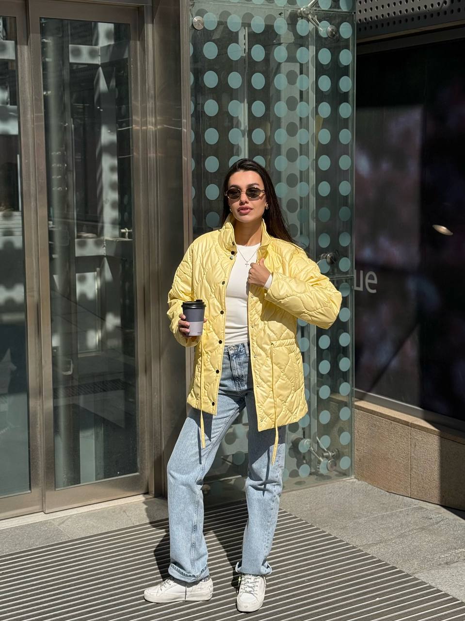 Person wearing a yellow jacket and sunglasses, holding a coffee cup, standing in front of a glass door.