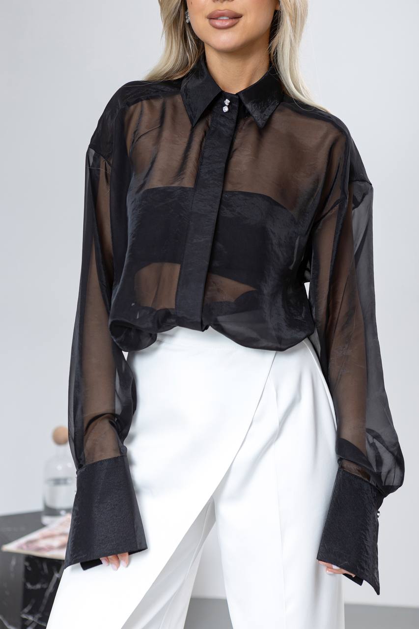 Person wearing a black sheer blouse over a black shirt with white pants on a plain background