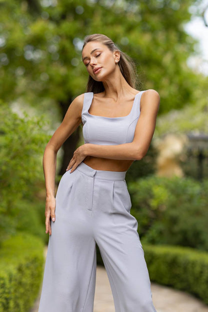 Woman in light gray outfit standing outdoors with greenery in the background