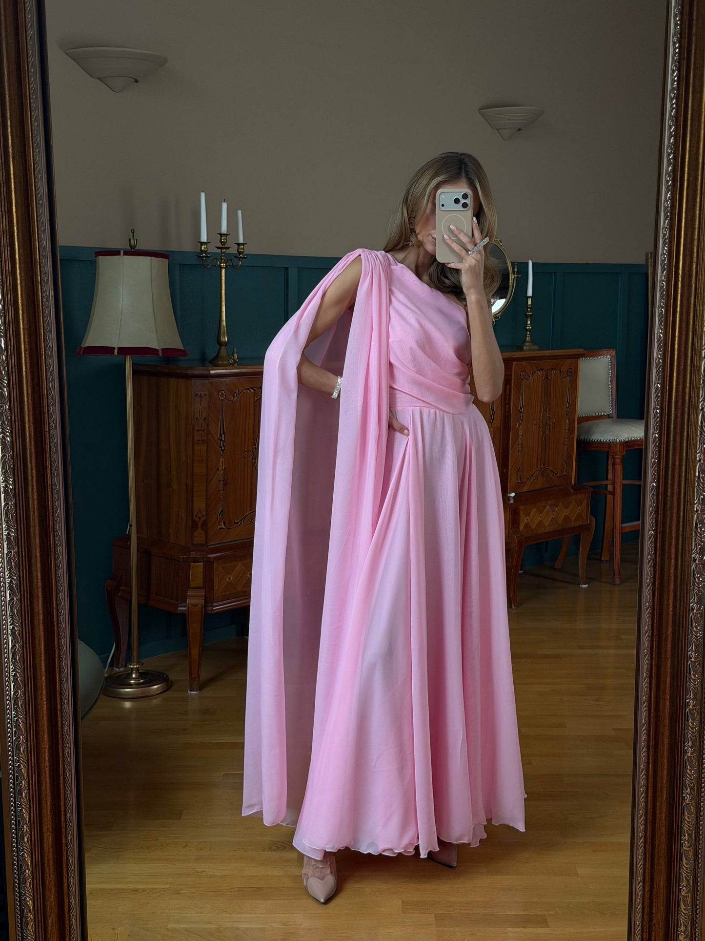 Person wearing a pink dress standing in a room with wooden furniture and a lamp.