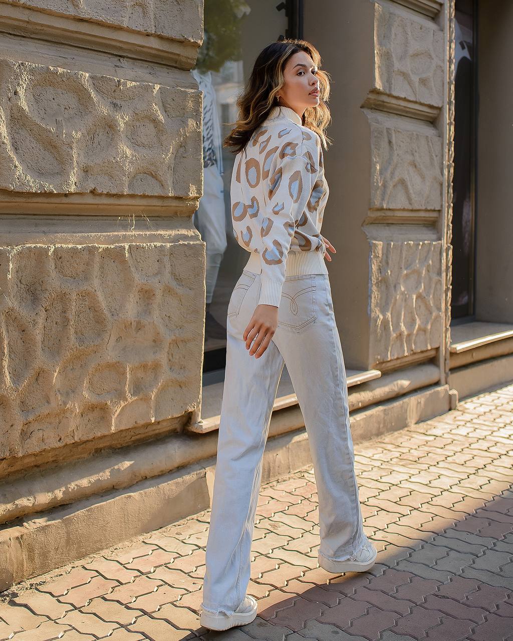 Woman in a leopard print top and light-colored pants standing on a stone pavement.