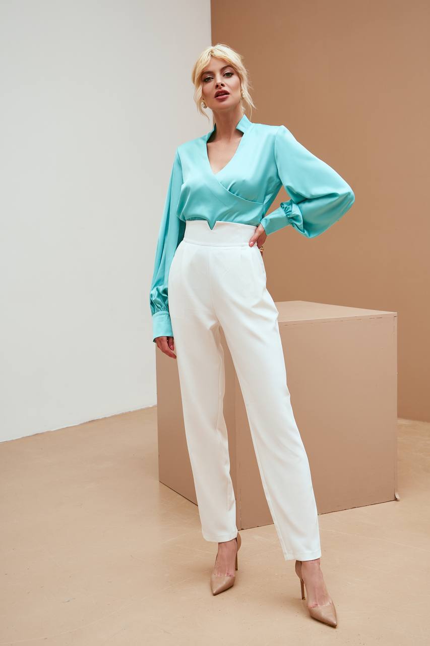 Woman wearing a light blue blouse and white pants standing against a beige background