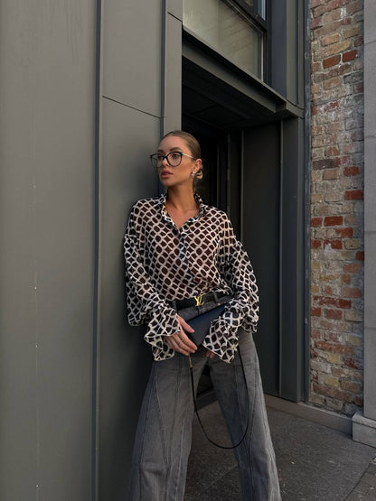 Woman in a patterned blouse and jeans leaning against a wall.