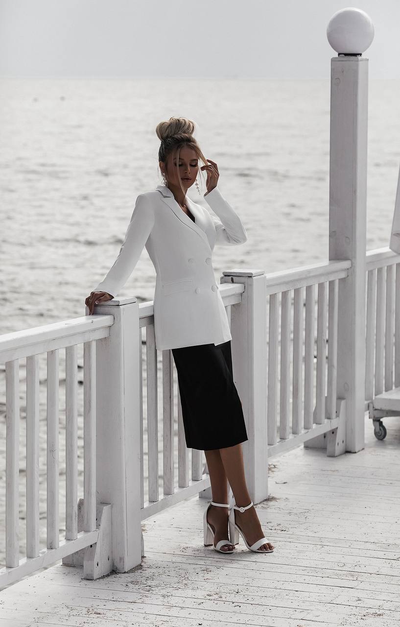 Woman in a white blazer and black skirt standing on a white wooden deck.