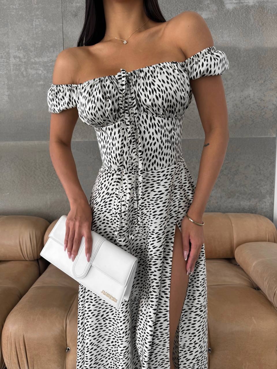 Woman wearing a leopard print off-shoulder dress holding a white clutch against a gray wall.