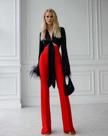 Woman wearing a black top and red pants with feathered accents against a white paneled wall.