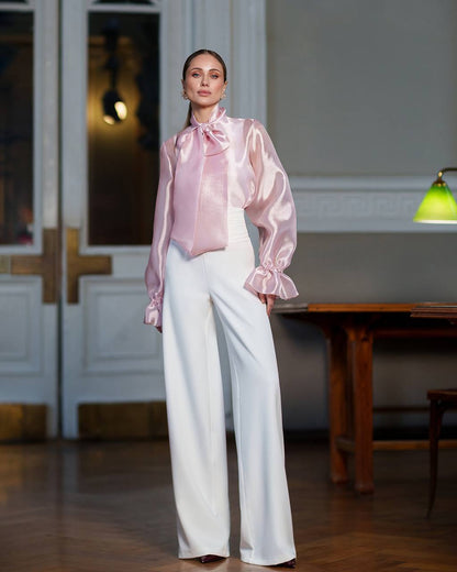 Woman wearing a pink blouse with a large bow and white pants in an indoor setting.