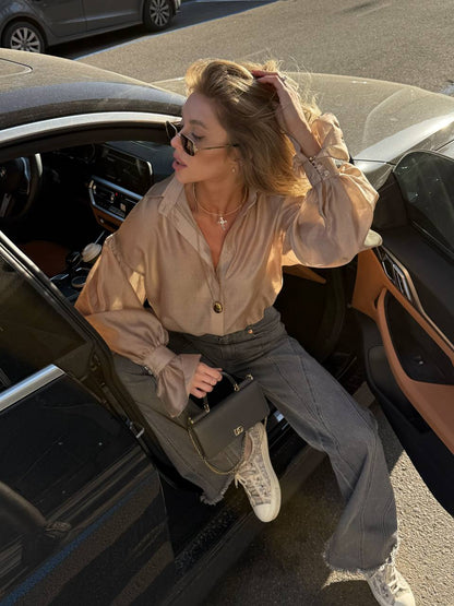 Woman sitting in a car wearing a beige blouse and gray pants, holding sunglasses.