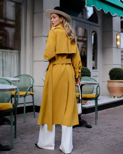 Woman in a yellow trench coat and white pants standing on a street with green chairs and tables.