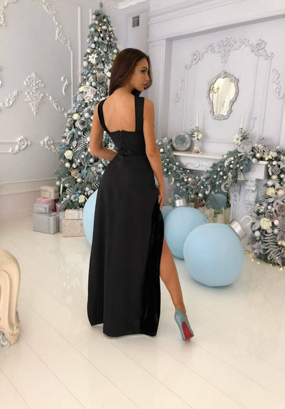 A woman wearing a sleeveless black floor-length evening gown with a high thigh slit, photographed in a decorated holiday-themed room with a Christmas tree, garlands, and light blue balloons.