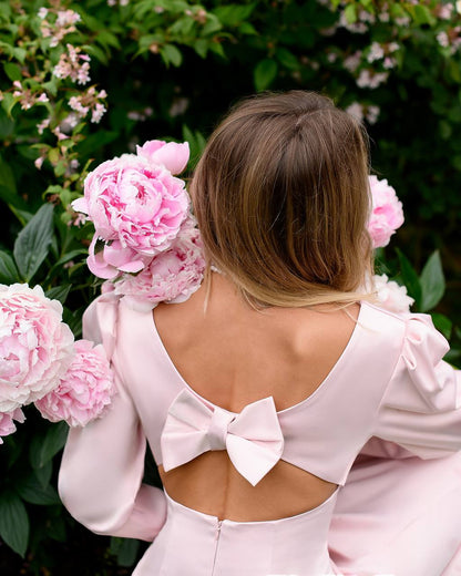 Woman in a pink dress with a bow detail standing among pink flowers.